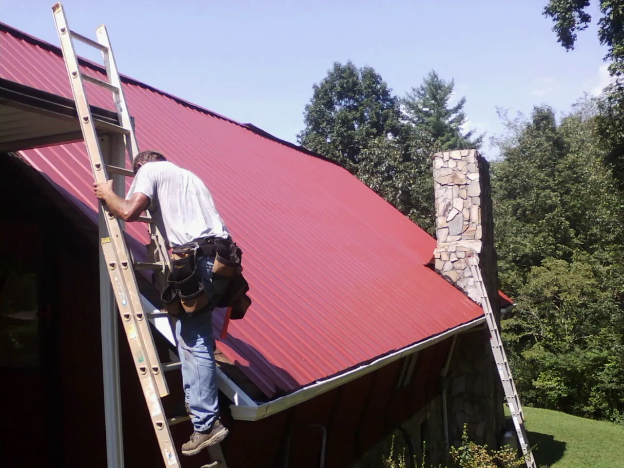 Roofer on a ladder beside a red metal roof and stone chimney — Soffit & Fascia Repair by Roofing Brothers in Boonsboro